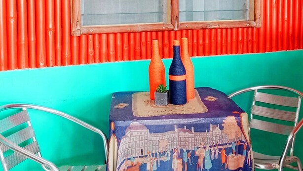 Una pequeña mesa con mantel colorido, dos sillas de metal y una ventana con persianas.