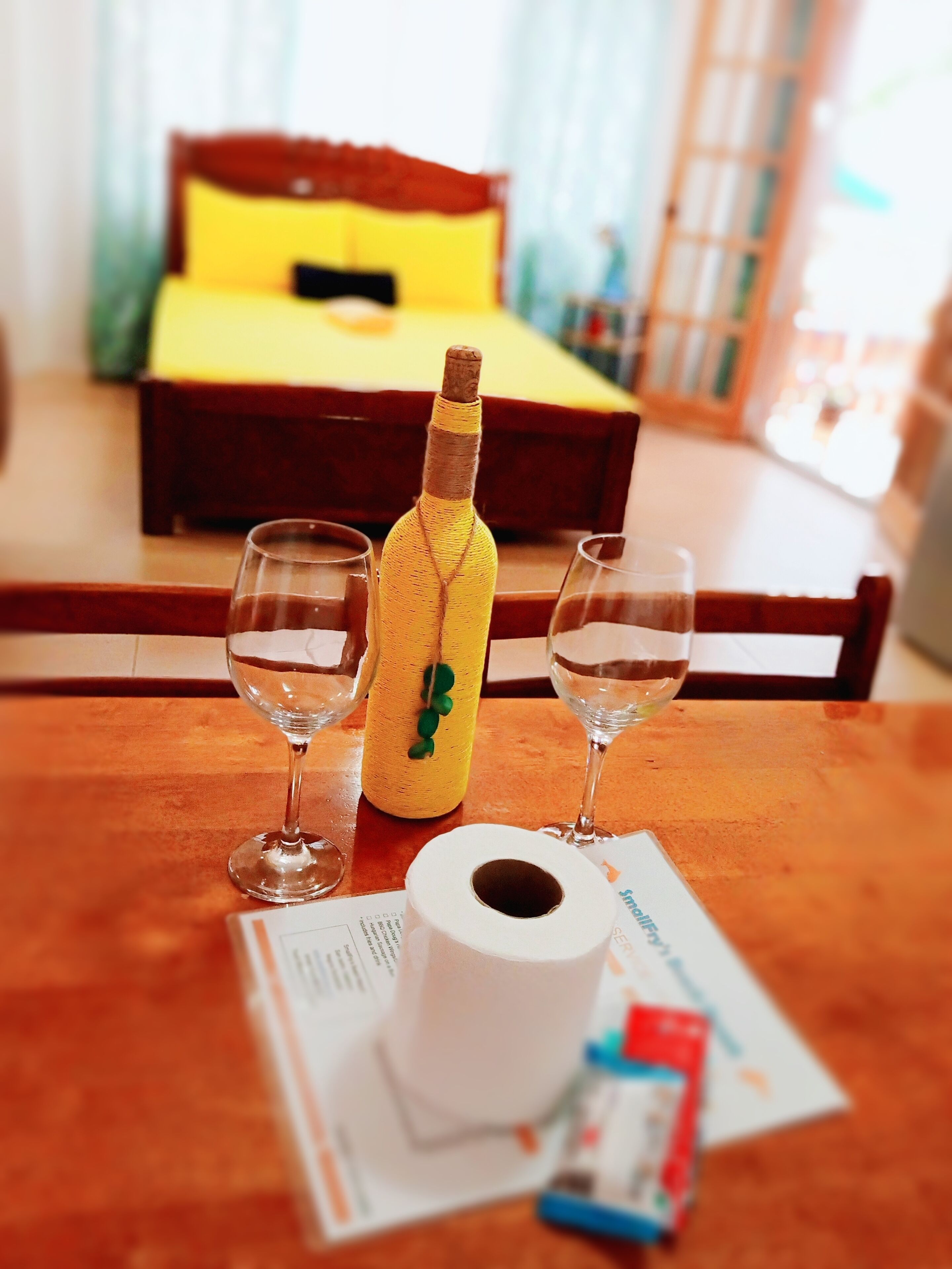 Camera d'albergo con un tavolo in legno, due bicchieri da vino, una bottiglia di vino, un rotolo di carta igienica e un letto con testiera gialla e biancheria rossa.