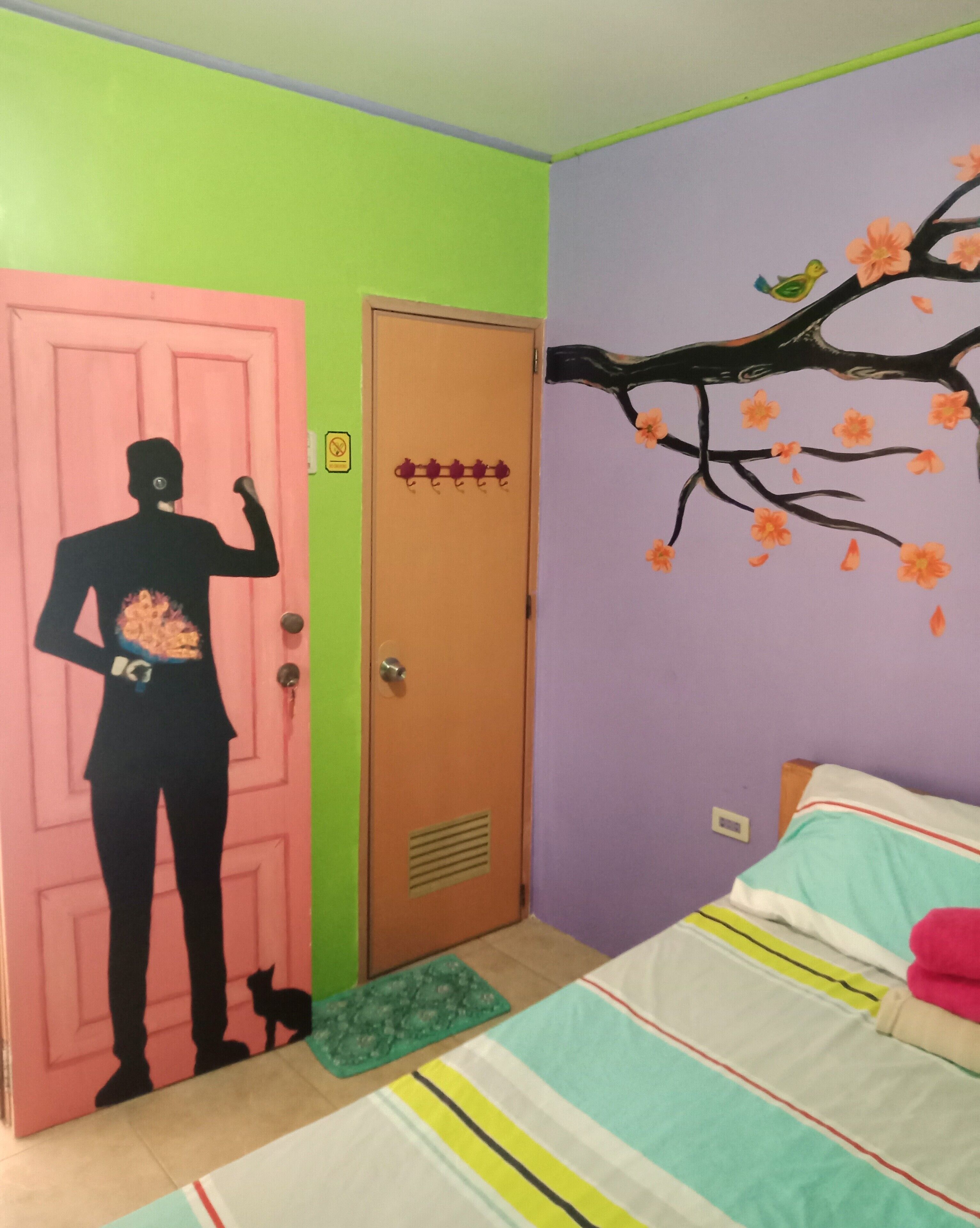 Camera da letto con una porta rosa, un letto con copriletto a righe, un murale raffigurante un albero fiorito e un piccolo tappeto sul pavimento.