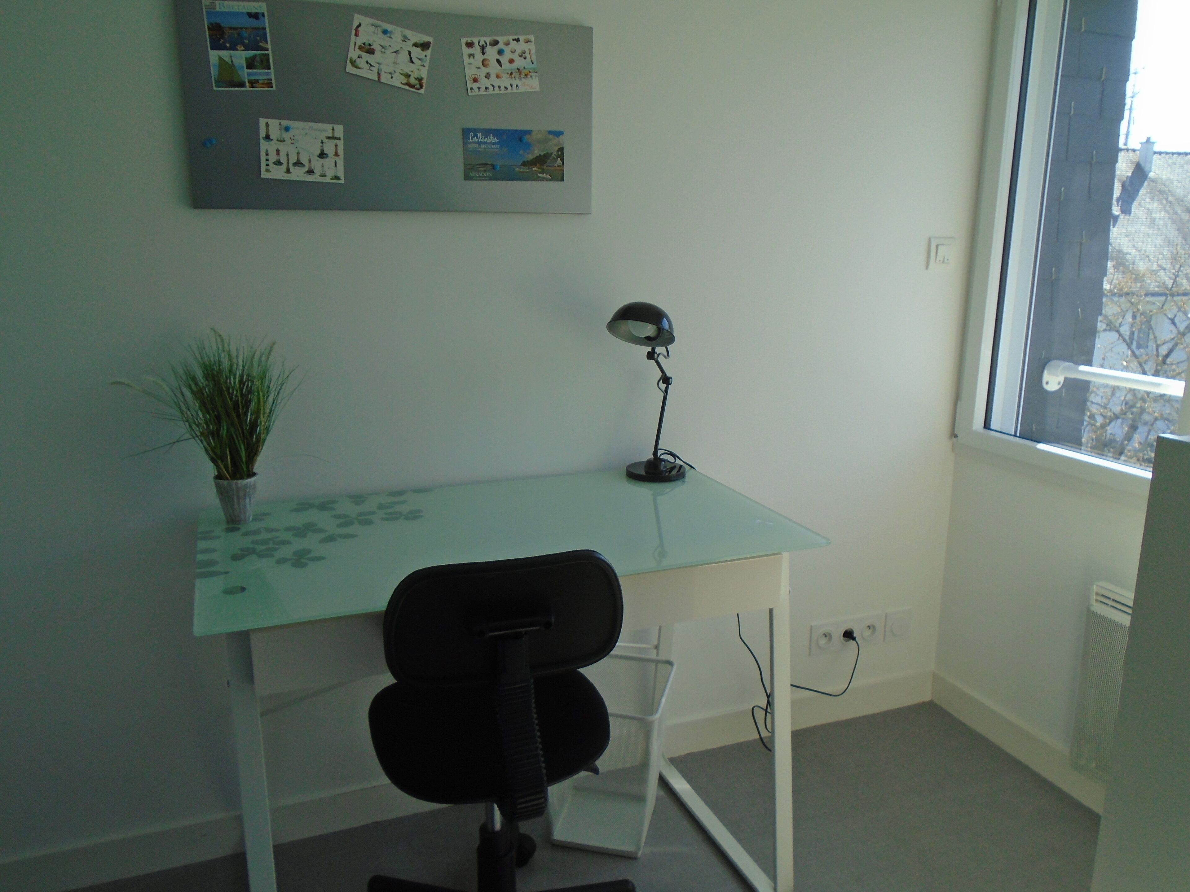 A small office with a glass desk, a black office chair, a potted plant, a desk lamp, and a bulletin board with various pictures and notes.