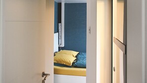 A narrow hallway leading to a bedroom with a bed, a wall-mounted heater, and a door with a handle.