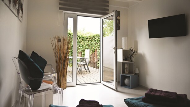 A room with a sliding glass door leading to an outdoor deck, a clear chair, a small table, and a bed with a blanket.