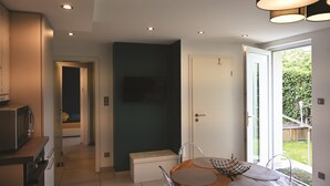 A modern kitchen with a microwave, a dining table with chairs, a television mounted on the wall, and a sliding glass door leading to a garden.