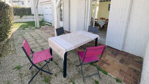 Une terrasse extérieure avec une table et deux chaises, un espace couvert avec une porte, et une vue sur l’intérieur de la maison.