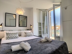 A modern bedroom with a large bed, two bedside tables, a ceiling light, and a window with a view of buildings.