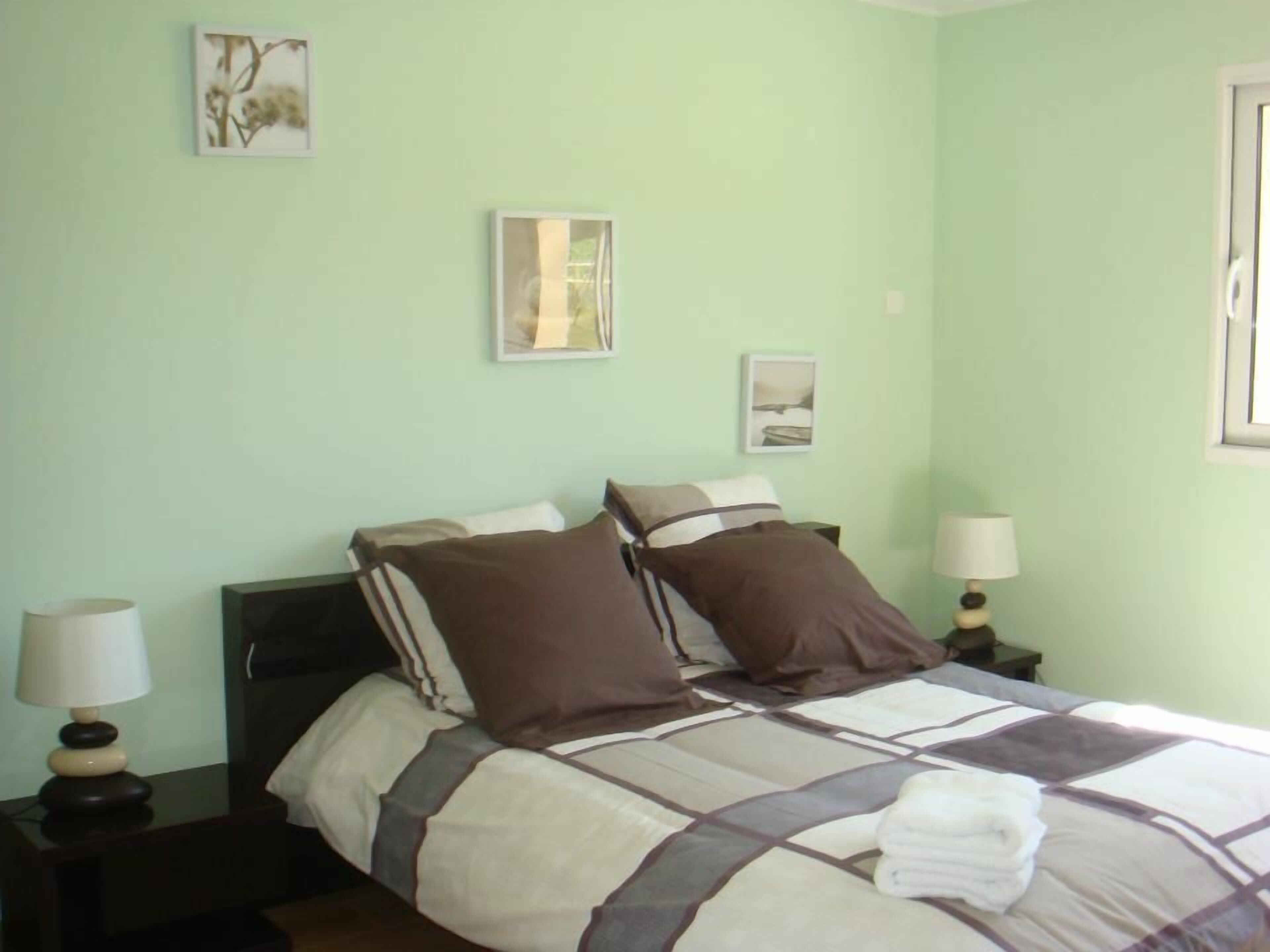 A neatly made bed with a checkered bedspread, two pillows, and a folded towel on the bed. There are two bedside tables with lamps, and the walls are painted light green.