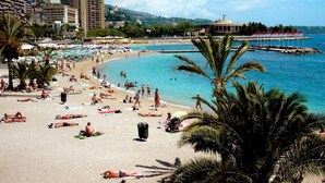 Une plage où des gens prennent des bains de soleil, une mer bleue et limpide, et des bâtiments en arrière-plan.