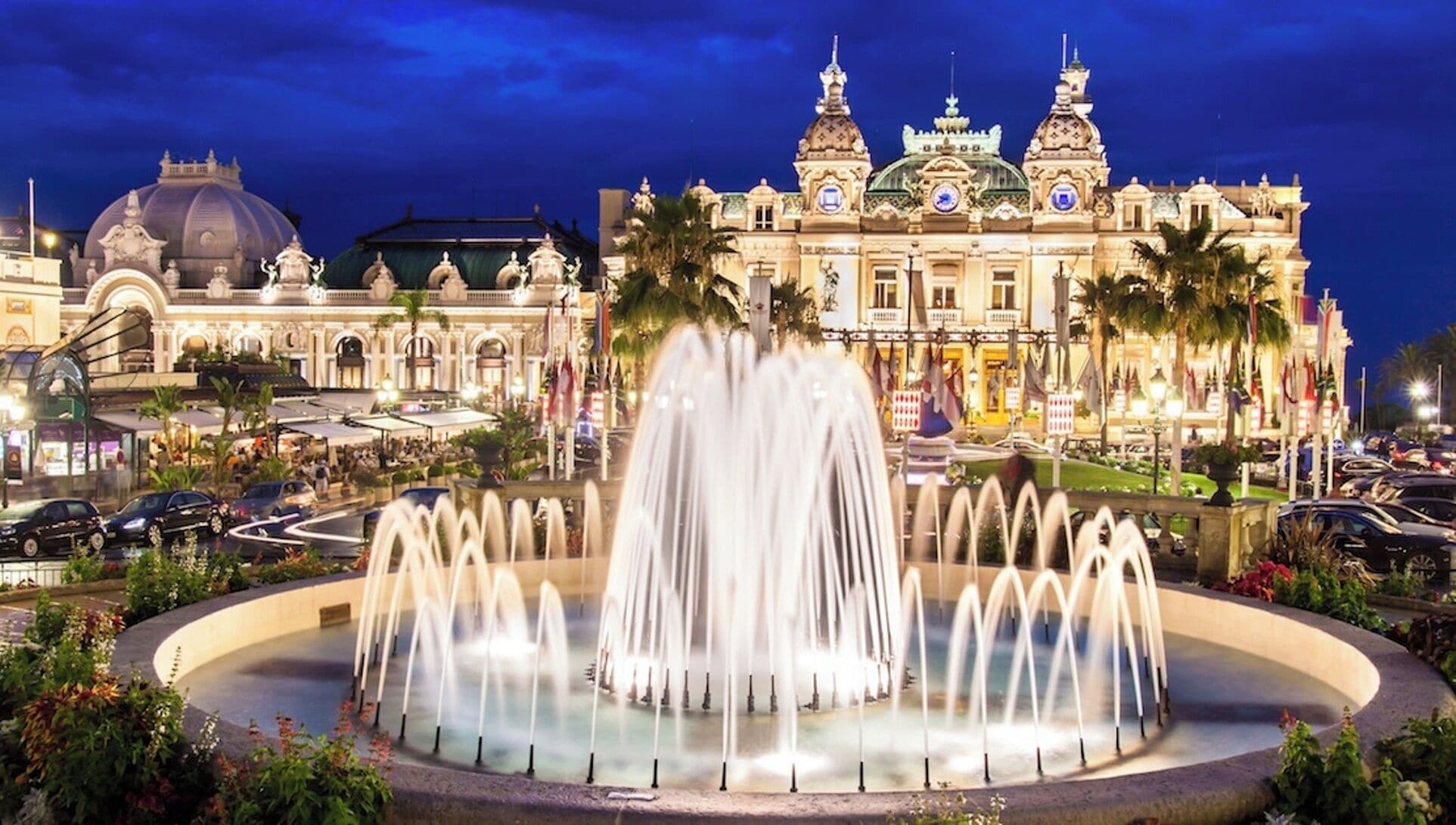 Un grand hôtel avec une vaste fontaine au centre, entouré de palmiers et d’une place bien éclairée.