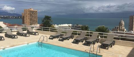 A rooftop pool with lounge chairs and a view of the sea and buildings.