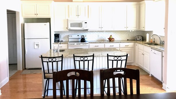 A modern kitchen with white cabinets, a central island with chairs, a microwave, a kettle, and a sink.