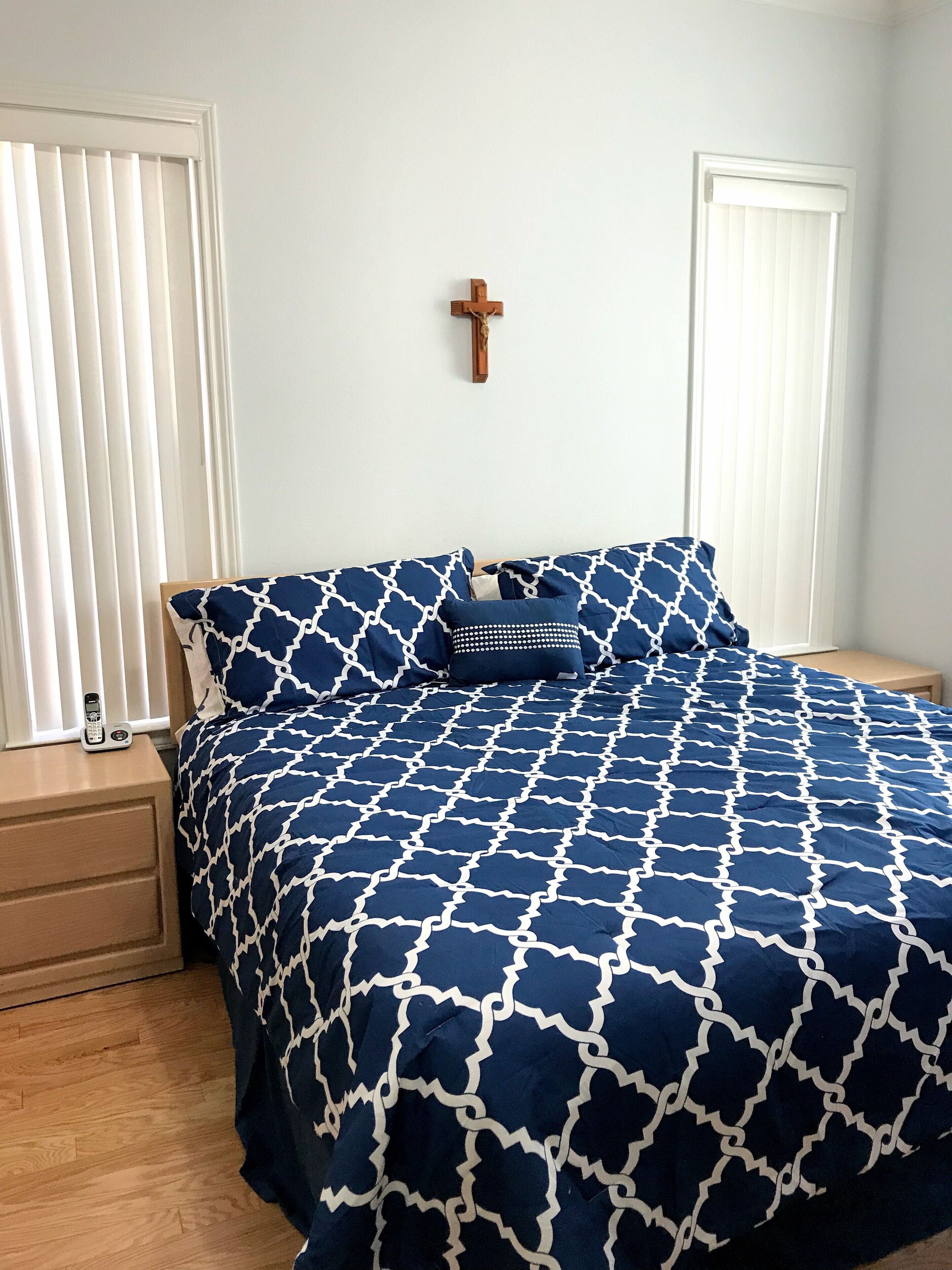 A bedroom with a bed covered in a blue and white patterned bedspread, a wooden nightstand with a telephone, and a wall-mounted cross.
