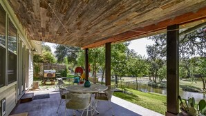 Un patio couvert comprenant une table et des chaises rondes en métal, un barbecue et une vue sur un parc avec un étang.