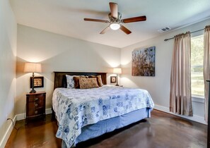A bedroom with a bed, two bedside lamps, a ceiling fan, a nightstand, a painting, and a window with curtains.