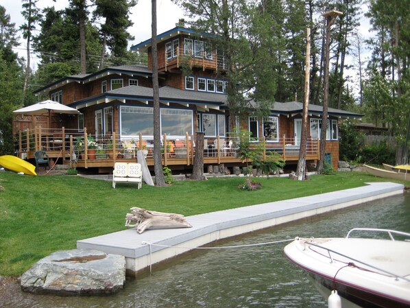 A two-story wooden house with a deck, a small dock, and a grassy yard.