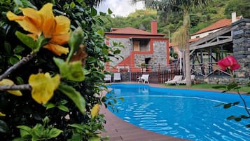 A swimming pool with a wooden deck, surrounded by lush greenery and a stone building with a red roof.