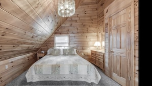 Ein gemütliches Schlafzimmer im Dachgeschoss mit Holzwänden, einem Bett mit floralem Bettzeug, einem Nachttisch, einer Kommode und einer Hängeleuchte.