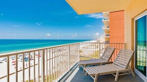 Een balkon met twee houten loungestoelen en een tafel, met uitzicht op een strand met een helderblauwe lucht en zee.