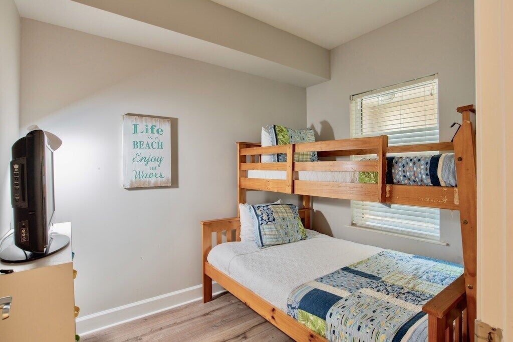 A bedroom with a bunk bed, a television on a stand, a window with blinds, and a motivational quote on the wall.
