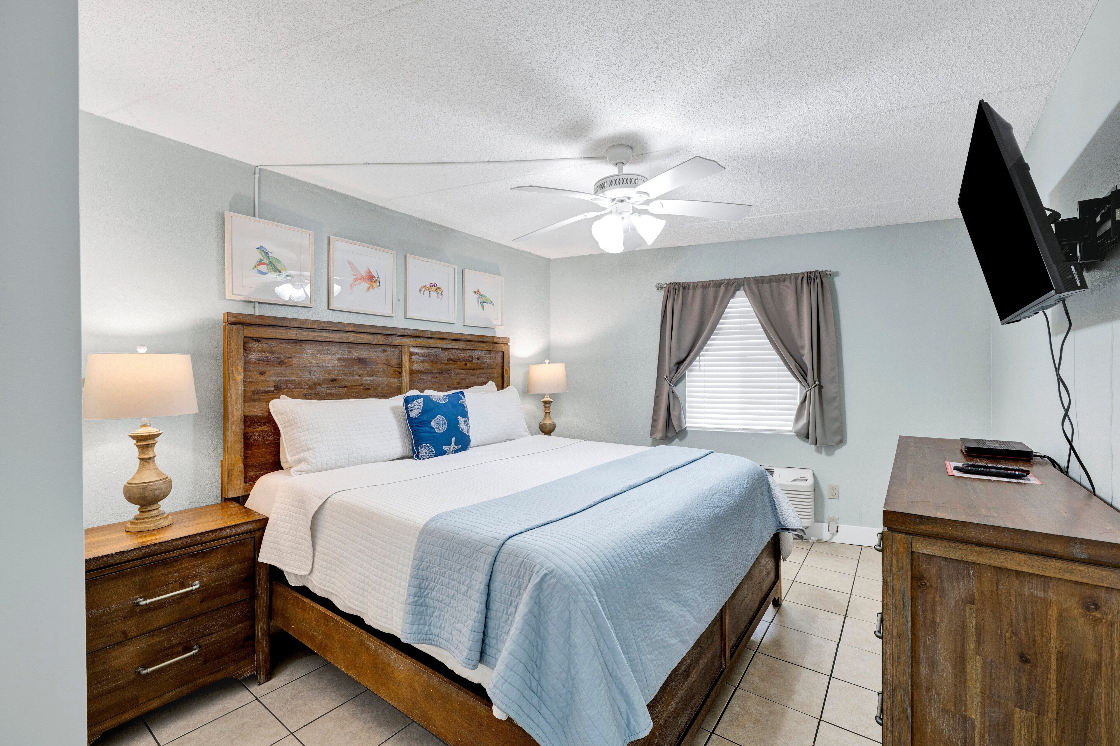 A bedroom with a wooden bed, two bedside tables with lamps, a ceiling fan, a flat-screen TV mounted on the wall, and a window with curtains.