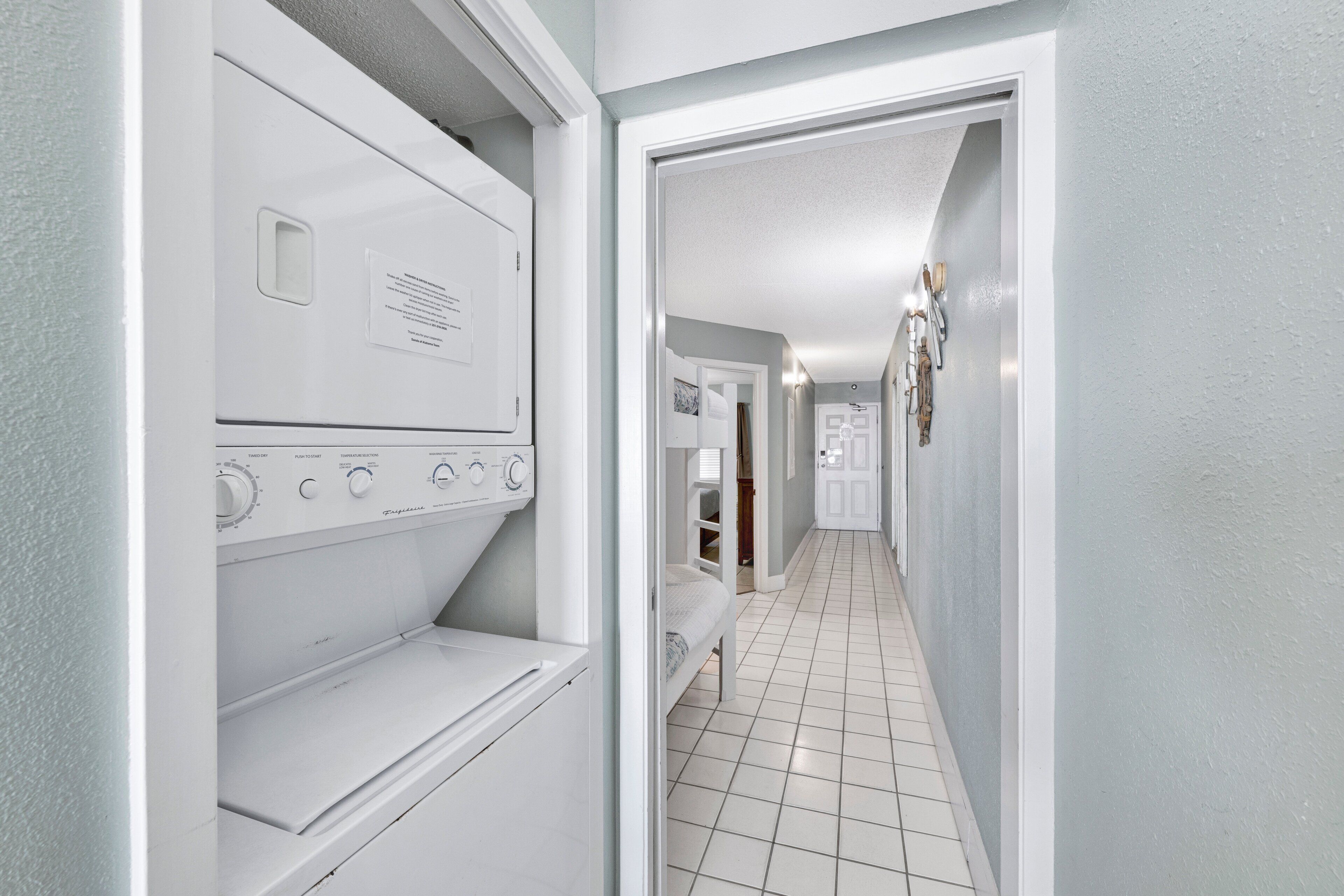 Une buanderie équipée d’un lave-linge et d’un sèche-linge à chargement frontal, d’un sol carrelé et d’un couloir menant à une chambre à coucher avec un lit et un placard.