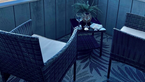 A balcony with wicker chairs and a table, overlooking a beach and residential area.
