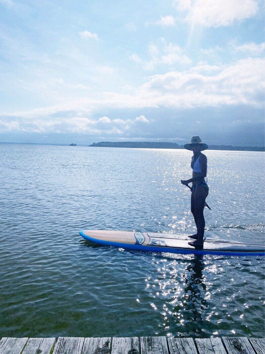 Een persoon staat op een paddleboard in rustig water, met een verre kustlijn en een bewolkte lucht.