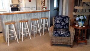 A kitchen with a bar area, white stools, a wicker chair, and a small table with a vase.