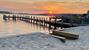A sandy beach with a wooden pier extending into calm water, a wooden bench, and a distant sunset.