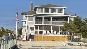 A two-story beachfront house with a balcony, a wooden deck, and a sandy beach in the foreground.
