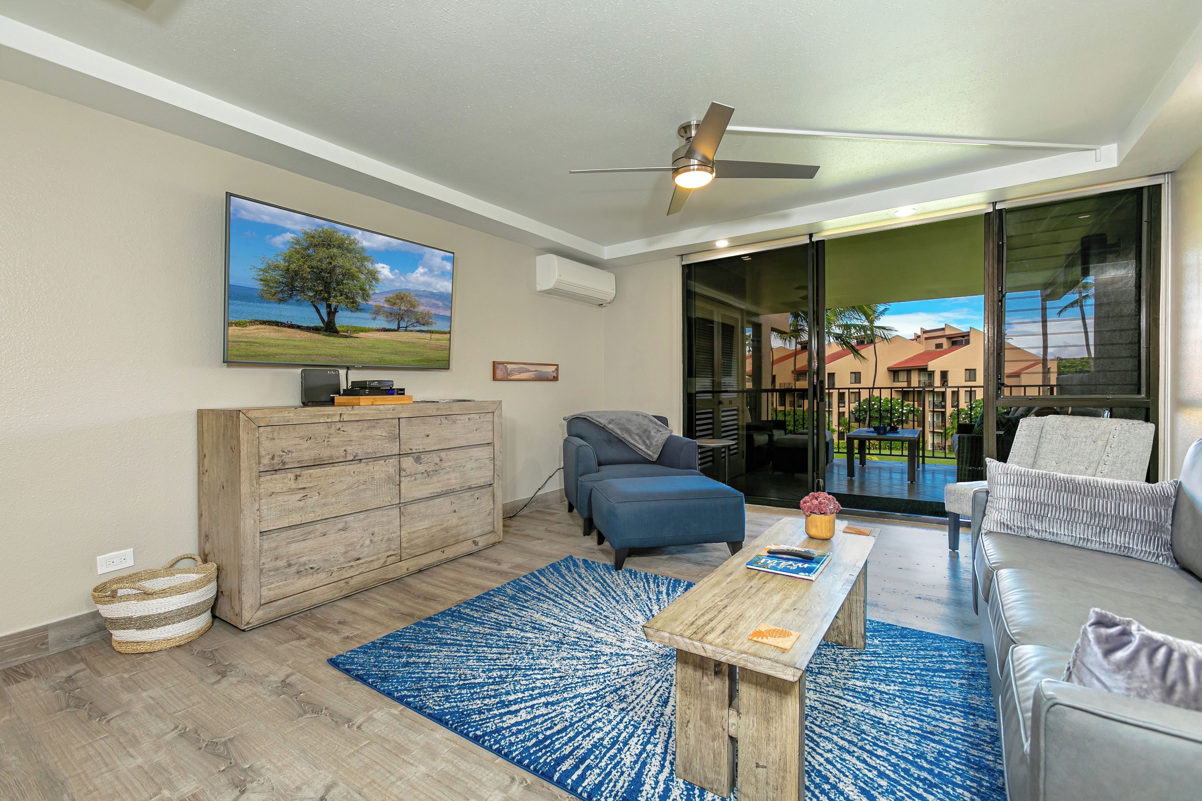 A modern living room with a flat-screen TV, wooden cabinet, blue and white rug, and a ceiling fan.