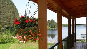 Een houten terras met een hangmand vol bloemen, uitzicht op een meer en een steiger met een boot.