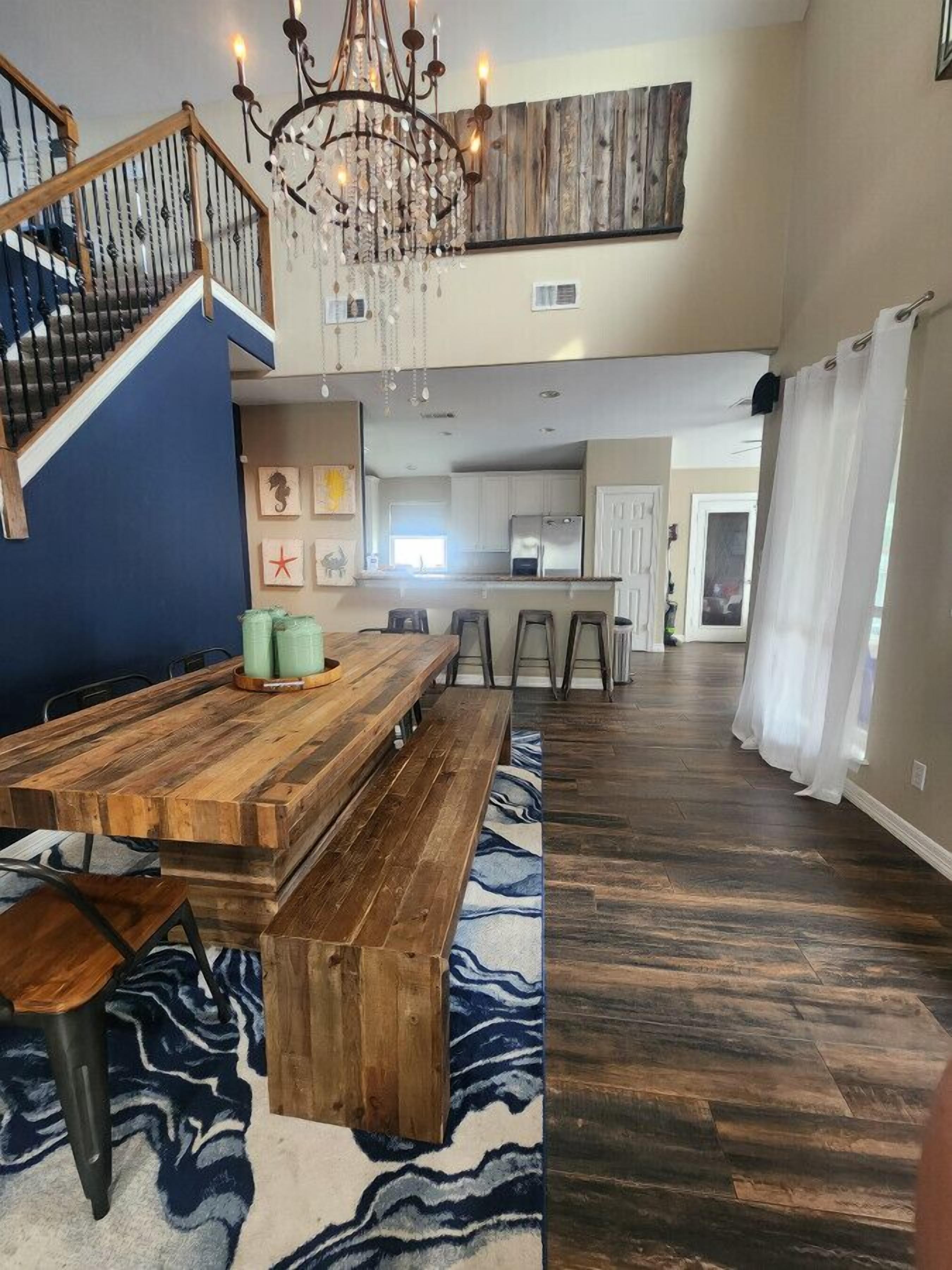 Un vaste espace dédié à la salle à manger, avec une table et des bancs en bois, un lustre, et une vue sur la cuisine et le salon.