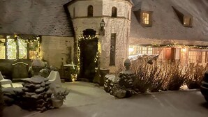 Ein schneebedecktes Haus mit einem kegelförmigen Dach, einem kleinen Turm und einer Haustür mit einem Kranz. Vor der Tür stehen Blumentöpfe und ein Stuhl.