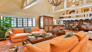 Ein geräumiges Wohnzimmer mit hohen Decken, großen Fenstern und einer gemütlichen Sitzecke mit orangefarbenen Sofas und einem Holztisch.
