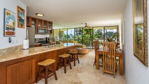 A spacious kitchen with a large island, wooden bar stools, and a dining area with a wooden table and chairs. - OCEANFRONT CONDO MAUI SUNSET#215-A, SLEEPS 4 (Kihei)