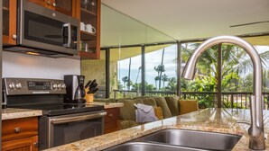 A modern kitchen with granite countertops, stainless steel appliances, and a large window overlooking a lush garden. - OCEANFRONT CONDO MAUI SUNSET#215-A, SLEEPS 4 (Kihei)