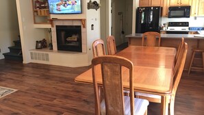 Un comedor con una mesa y sillas de madera, un televisor sobre la chimenea, y una cocina con refrigerador y microondas.
