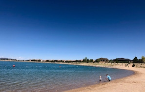 Ein Strand mit einem klaren, blauen See, vereinzelten roten Bojen und einigen Personen. Der Text 'Lake at the Hill' ist sichtbar.