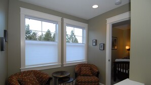 A room with two patterned armchairs, a small round table, and a white bench. There are two large windows with blinds, a door leading to another room, and a ceiling light.