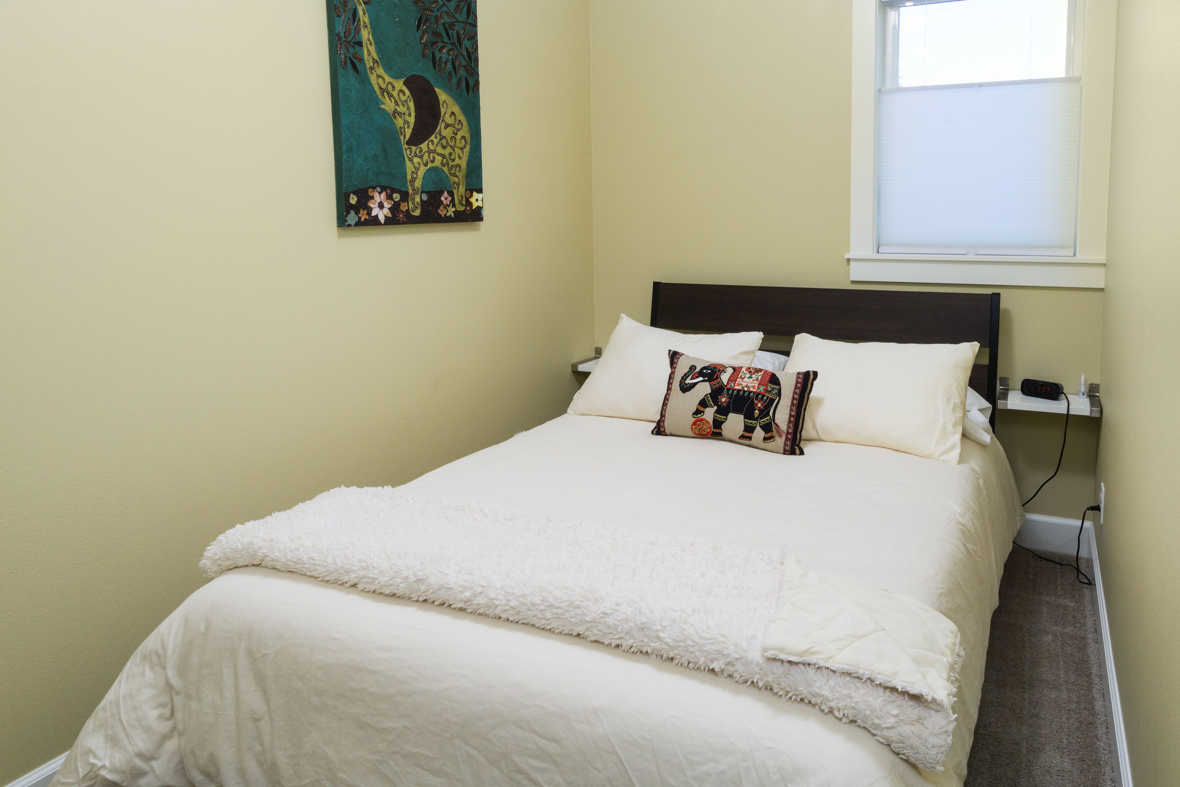 A neatly made bed with white bedding and two decorative pillows, a wall hanging with a giraffe design, and a window with blinds.