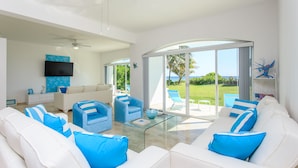 A spacious living room with white and blue furniture, a glass coffee table, and a large sliding glass door leading to an outdoor area with a view of the ocean. - Views, Views, Views!! (Puerto Morelos)
