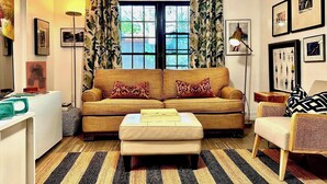 A cozy living room with a sofa, ottoman, and armchair, a window with curtains, and framed pictures on the walls. - Tropical, private, downtown (Sarasota)