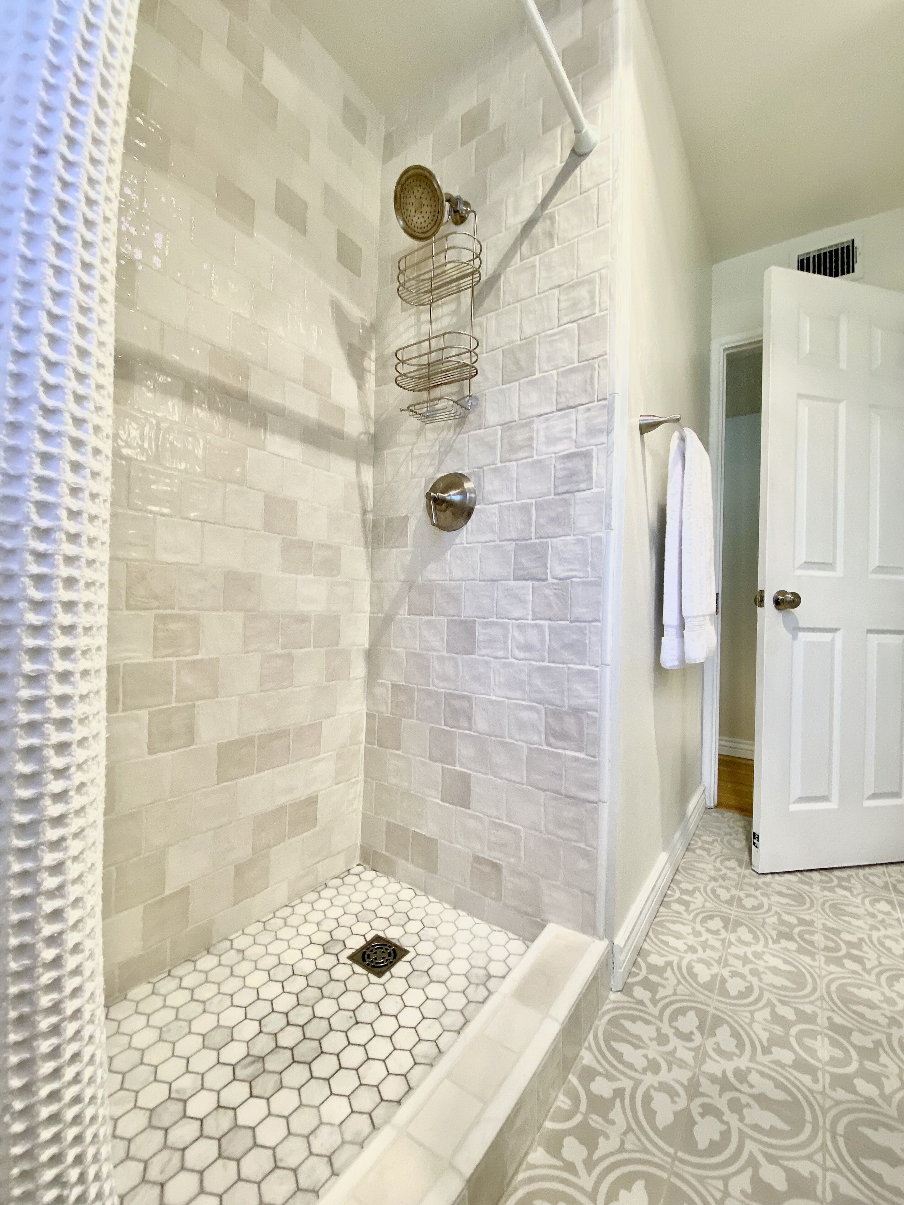 A bathroom with a shower, tiled walls, a white door, and a patterned floor.