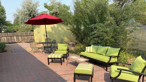 Un espace extérieur aménagé en patio, avec un sol en briques, des meubles rembourrés de couleur verte et un parasol rouge.