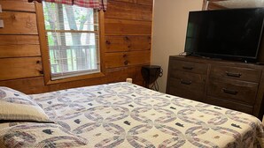 A bedroom with a wooden wall, a bed with a patterned quilt, a TV on a wooden dresser, and a window with blinds.