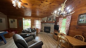 A cozy cabin interior with a fireplace, leather sofa, wooden dining table, and a ceiling chandelier.