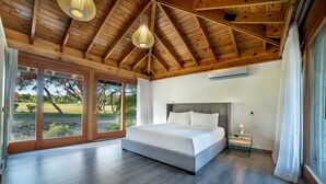 Ein geräumiges Schlafzimmer mit einem großen Bett, Holzbalkendecke und Blick auf einen Golfplatz durch Glastüren.
