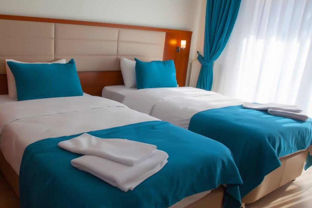 A hotel room with two beds, each with a blue bedspread and white pillows. There is a nightstand with a lamp, and the room has a wooden headboard.