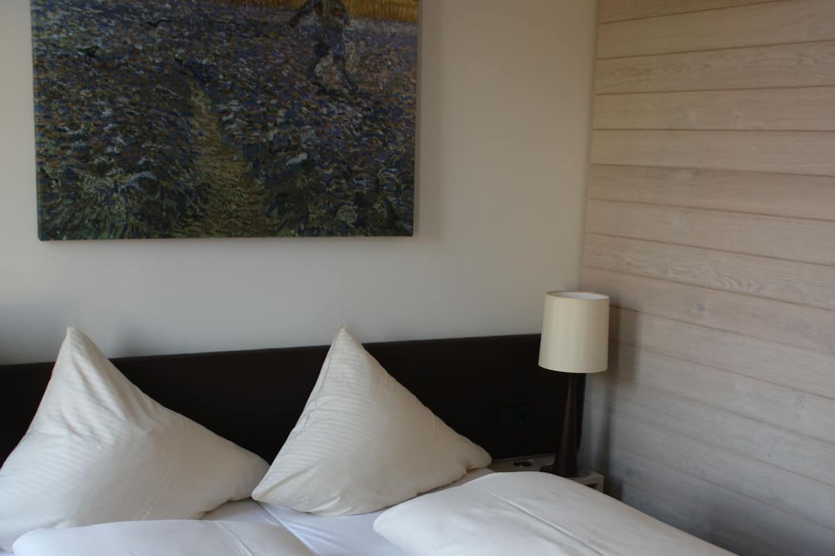 A double bed with white linens and pillows, a bedside table with a lamp, and a painting of a field with a person on the wall.