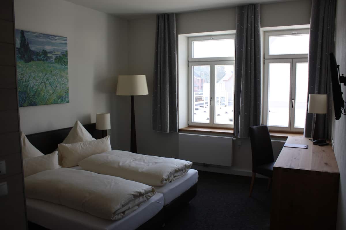A hotel room with two beds, a desk, a chair, a lamp, a painting, and a window with curtains.